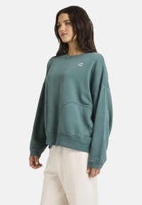 Sudadera grande de color verde azulado en tejido suave y texturizado, con un escote redondo, puños acanalados y bolsillos laterales. Presenta un pequeño logo en el pecho.