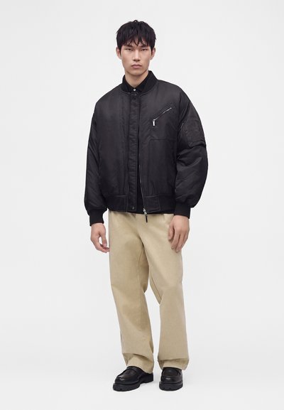 Άνδρας με μαύρο bomber jacket, μπεζ ευρύχωρο παντελόνι και μαύρα παπούτσια, στέκεται μπροστά σε απλό φωτεινό φόντο.