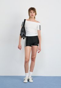 Balti off-the-shoulder marškiniai su juodomis juostelėmis, derinami su juodomis šortais, kuriuose yra trys juostelės, ir baltomis sportinėmis bateliais. Juoda rankinė laikoma rankoje.