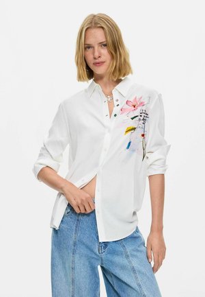 Blonde vrouw draagt een losse witte blouse met knopen en bloemen- en abstracte ontwerpen, gecombineerd met lichtblauwe spijkerbroek, poserend tegen een witte achtergrond.
