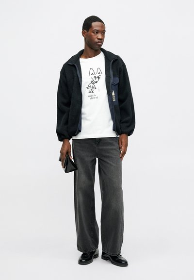 Μαύρο φλις μπουφάν, λευκό μπλουζάκι με γραφικό σχέδιο και καρτούν, γκρι φαρδιά παντελόνια και μαύρα παπούτσια. Μοντέλο που κρατά μια μαύρη πορτοφόλι.