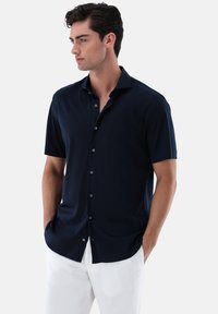 Marineblaue kurzärmelige Hemd aus glattem Stoff mit einer geknöpften Leiste und einem flachen Kragen, getragen zu weißen Hosen.