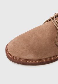 Close-up van de tan suede leren schoentip met een bruine gestikte zool en zichtbare veters op een witte achtergrond.
