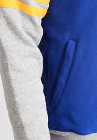 Sudadera azul y gris con un bolsillo lateral, con mangas grises y rayas amarillas en la parte superior del brazo, hecha de una tela suave.