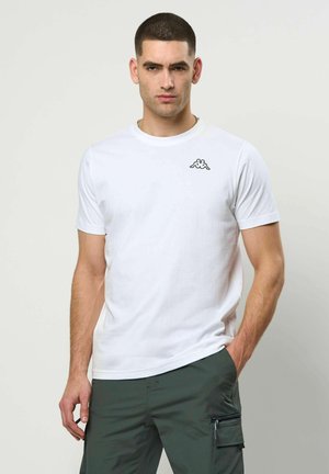 Młody mężczyzna o krótkich włosach, ubrany w białą koszulkę z logo i ciemnozielone spodnie typu cargo, stoi z jedną ręką w kieszeni.