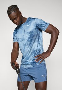 Blauwe sport T-shirt met een gestructureerd ontwerp, voorzien van een wit logo. Gecombineerd met lichtblauwe shorts, met een zwart handvat.