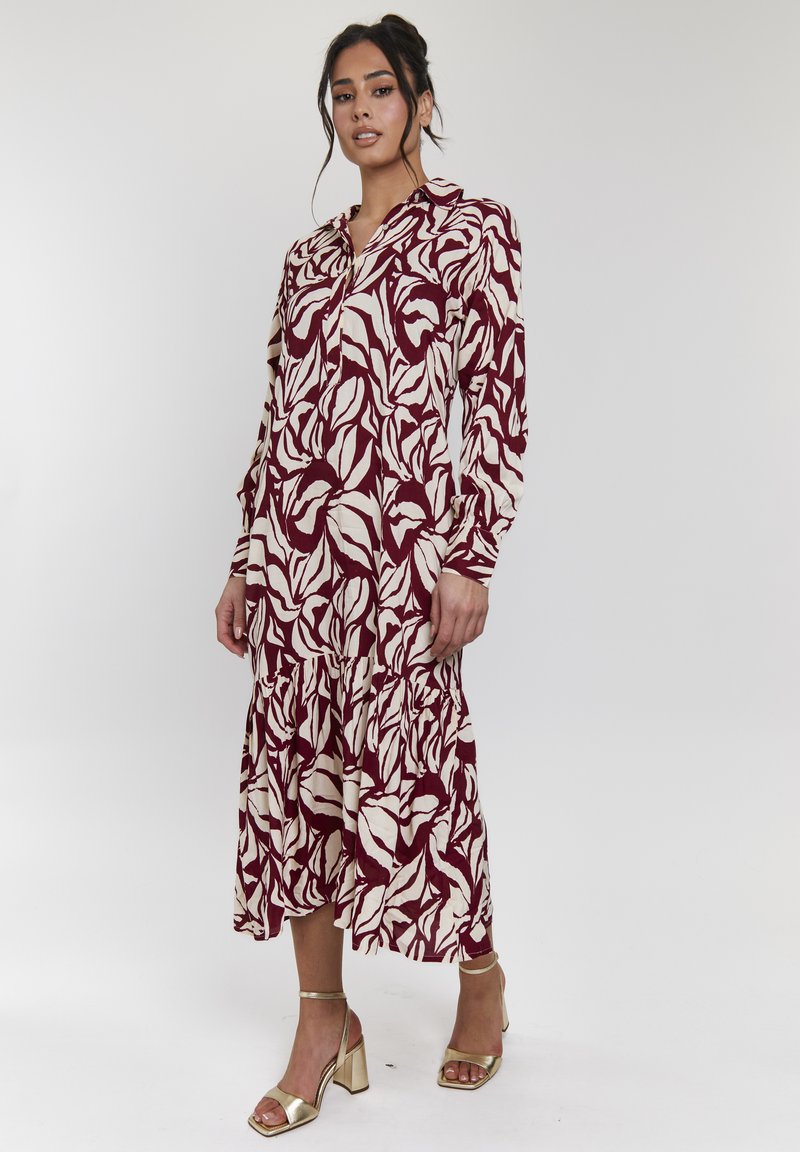 Langes, kastanienbraunes Kleid mit weißen Blattmustern, knopfbar vorne und mit Rüsche am Saum. Kombiniert mit goldenen, metallischen Absatzsandalen mit Riemen. Glatte Stoffoberfläche.