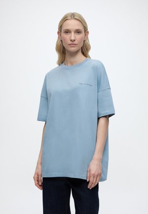 Γυναίκα με ξανθά μαλλιά που φοράει ανοιχτό μπλε χαλαρό μπλουζάκι με το κείμενο "keep on moving" και σκούρο παντελόνι, στέκεται μπροστά σε ένα απλό φόντο.