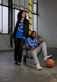 Du modeli sportiniu apranga, vienas vilkintis mėlyną marškinėlį su užrašu 'BUCKS' ir pilkas kelnes, kitas vilkintis juodą švarką ir mėlyną marškinėlį. Treniruotės aplinka, krepšinis.