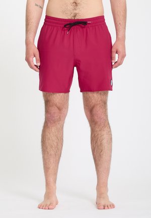 Homme portant un short de bain bordeaux mi-cuisses avec un cordon noir, debout pieds nus contre un fond blanc uni.
