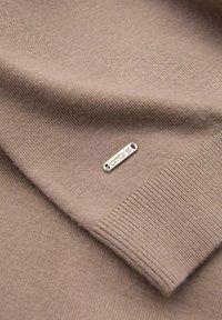 Beiger Pullover aus weichem Stoff, mit einem gerippten Saum und einem kleinen silbernen Etikett mit dem eingeprägten Schriftzug "ombre". Glatte Textur und minimalistisches Design.