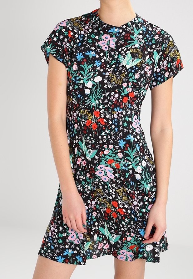 Femme portant une robe noire à manches courtes avec un motif floral et d'oiseaux coloré, se tenant devant un fond clair uni.