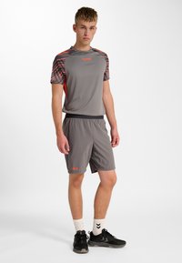 Grå atletisk tröja med mönstrade axlar och orange accenter, kombinerad med grå shorts. Bärs med svarta skor och vita strumpor.