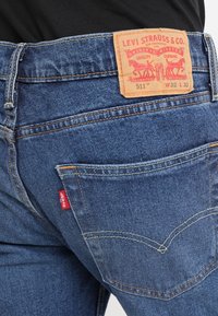 Jeans de denim azul ajustados con costuras visibles, una etiqueta de cuero en la cinturilla y una pequeña etiqueta de logo roja en el bolsillo trasero.