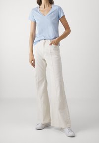 Ljusblå V-ringad t-shirt med en liten logo, i kombination med vita vida byxor. Modellen bär vita sneakers. Minimalistisk design, slät textur.