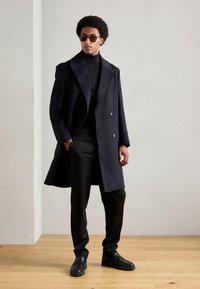 Marineblauwe wollen dubbelzijdige jas met geknopt revers, gecombineerd met een zwarte coltrui, zwarte broek en elegante zwarte schoenen.