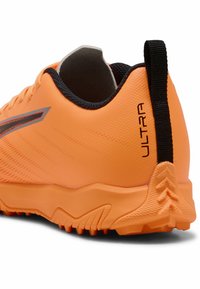 Zapatilla deportiva naranja con un upper suave, detalles en negro y una suela texturizada con patrones de agarre distintivos. "ULTRA" está impreso en la parte trasera.