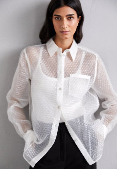 Camicia trasparente a bolle di colore bianco, con colletto, taschino frontale e chiusura con bottoni, abbinata a pantaloni scuri.