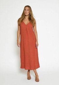 Femme aux cheveux longs portant une robe midi sans manches de couleur rouille et des sandales beiges à bout ouvert, se tenant devant un fond blanc.