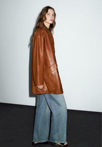 Bruine leren oversized jas, met een kraag en knoopsluiting. Gecombineerd met lichte blauwe denim jeans met een wijde pijp.
