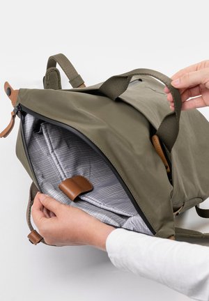 Zaino verde oliva con dettagli in pelle marrone, dotato di apertura con zip e di un interno grigio a quadretti. Le maniglie sono imbottite per maggiore comfort.