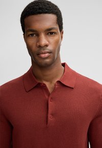 Man met kort krullend haar die een rustkleurige gebreide polo draagt, recht vooruit kijkend tegen een effen lichtgrijze achtergrond.