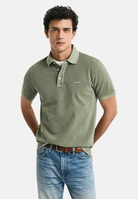 Olivgrünes Polo-Shirt mit kurzen Ärmeln, drei Knopfverschluss und dezentem Logo. Kombiniert mit hellblauen Denim-Jeans und braudem Gürtel.