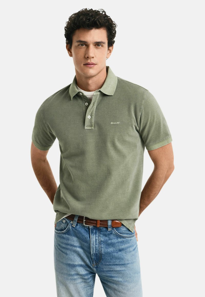 Olivgrünes Polo-Shirt mit kurzen Ärmeln, drei Knopfverschluss und dezentem Logo. Kombiniert mit hellblauen Denim-Jeans und braudem Gürtel.