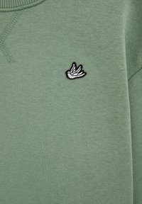 Vyšívaný biely vták na svetlozelenom plátne s viditeľným prešívaním pri ľavom hornom rohu.