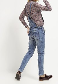 Salopette en denim bleu clair avec des bretelles ajustables et une coupe décontractée. Comprend des poches arrière et un léger délavage du tissu. Associée à des chaussures foncées.