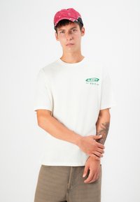 Cream-colored cotton t-shirt featuring green logo print on the left chest, paired with a pink and black cap. Model has visible arm tattoo.