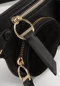 Černá kožená taška se zlatými zipy, kulatou semišovou kapsou, hladkým povrchem a kroužky na klíče. Funkční design s kontrastními materiály.