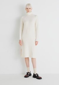 Rochie tricotată crem cu guler înalt, mâneci lungi, textură ribbed, fante laterale și pantofi cu platformă negri, în contrast, purtați cu șosete până la glezne.