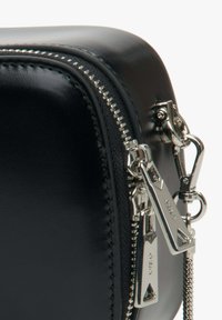 Zwarte leren tas met een glanzende afwerking, voorzien van een zilveren rits en hardware. De rits is voorzien van een geometrisch ontwerp en een afneembare ketting.