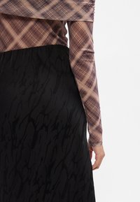 Camisa de manga comprida em tecido xadrez castanho, combinada com uma saia preta fluida com um padrão texturizado. Primeiro plano da parte superior do corpo e da mão.