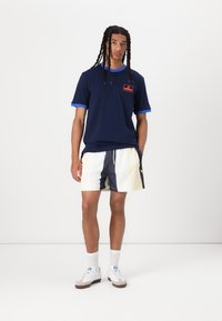 Mörkblå t-shirt med blå kant och röd logotyp, i kombination med randiga shorts i vitt och marinblått. Vita sneakers med grå detaljer och ankelsockor.