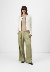 Giacca lavorata a maglia bianca con rifiniture nere, top beige e pantaloni di raso verde oliva larghi. La modella indossa occhiali da sole rossi e scarpe nere.