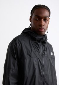 Veste à capuche noire en matériau léger, dotée d'une fermeture éclair intégrale et d'un logo triangulaire sur la zone gauche de la poitrine, avec une texture lisse.