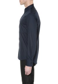 Homme portant une chemise ajustée bleu foncé à manches longues boutonnée et un pantalon noir, debout de profil sur fond blanc.