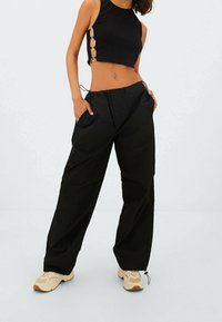 Zwarte cropped tanktop met zijuitsneden, gecombineerd met losse zwarte broek met een elastische tailleband en trekkoord, afgemaakt met beige sneakers.