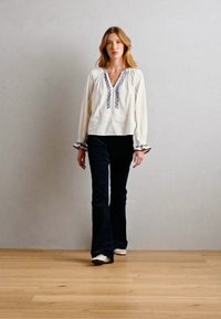 Weißes Blusenshirt mit marineblauer Stickerei, ausgestattet mit einem V-Ausschnitt und luftigen Ärmeln; kombiniert mit dunklen Schlaghosen und weißen Schuhen. Hintergrund aus Holzboden.