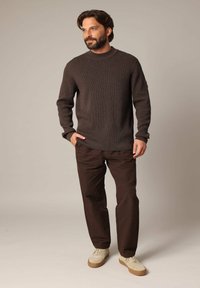 Un pull en maille marron foncé avec un col montant, associé à un pantalon marron ample et des baskets claires, sur un fond neutre.