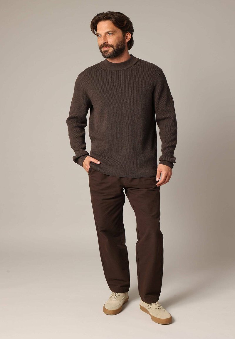 Un pull en maille marron foncé avec un col montant, associé à un pantalon marron ample et des baskets claires, sur un fond neutre.