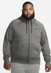 Šedá zipsová mikina s kapucňou, raglánovými rukávmi, prednými vreckami a čiernym logom Nike na hrudi. Vyrobené z textúrovanej tkaniny.