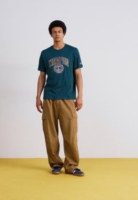 Camiseta de algodão verde-água com o logótipo "Champion", combinada com calças de carga beje com múltiplos bolsos, usadas com sapatos azuis e brancos numa superfície amarela.