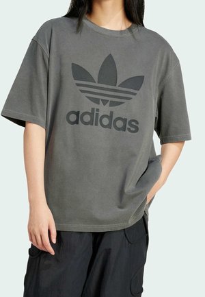 Henkilö, joka pukeutunut löysään harmaaseen Adidas-T-paitaan, jossa musta logo ja teksti, yhdistettynä mustiin cargo-housuihin, käsi taskussa, vaaleaa taustaa vasten.