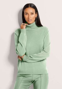 Frau mit langen dunklen Haaren, die einen hellgrünen Rollkragenpullover und passende Hosen trägt, posiert mit der Hand nah am Gesicht vor einem weißen Hintergrund.