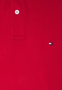 Červené polo tričko s texturovanou tkaninou, tříknoflíkovým zapínáním s bílými knoflíky a malým vyšitým logem v červené, bílé a modré barvě.