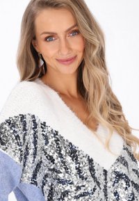 Sweter typu V-neck w białym i jasnoniebieskim kolorze, ozdobiony cekinowym wzorem z srebrnymi i niebieskimi akcentami. Miękka tekstura, luźny krój i casualowy styl.