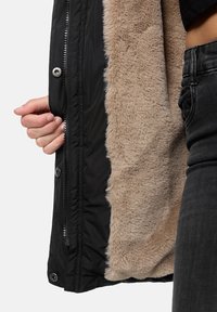 Svart ytterjacka med en mjuk, beige fuskpälsfoder. Har silverfärgade detaljer och dragkedjestängning, visad med en hand som håller i dragkedjan.